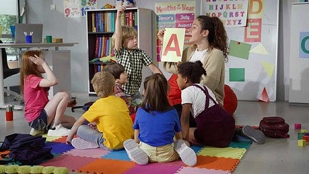 a teacher holds up a paper with the letter A on it while preschoolers gather around